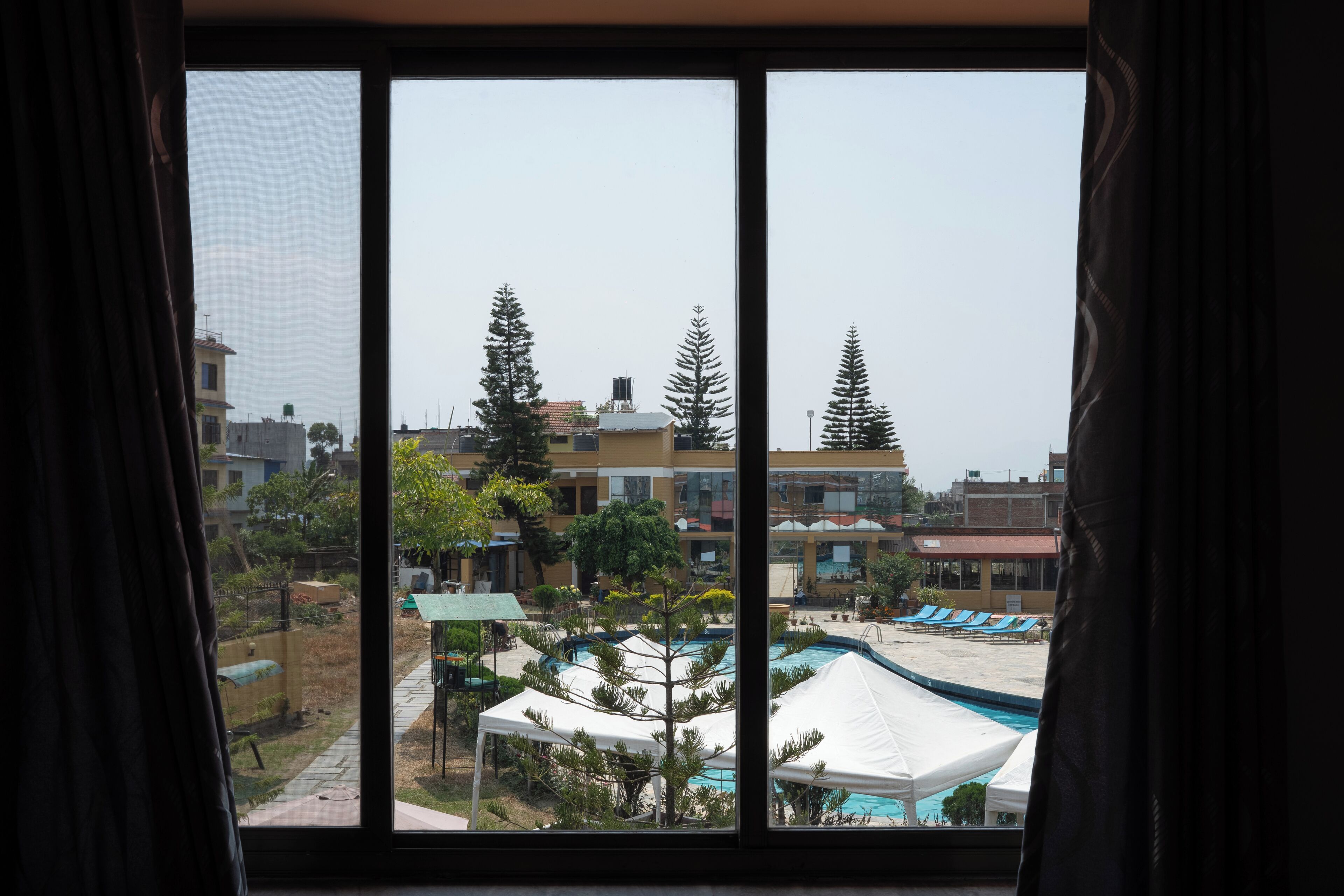 Vista a través de una ventana de un hotel con piscina, sillones de descanso y una carpa.
