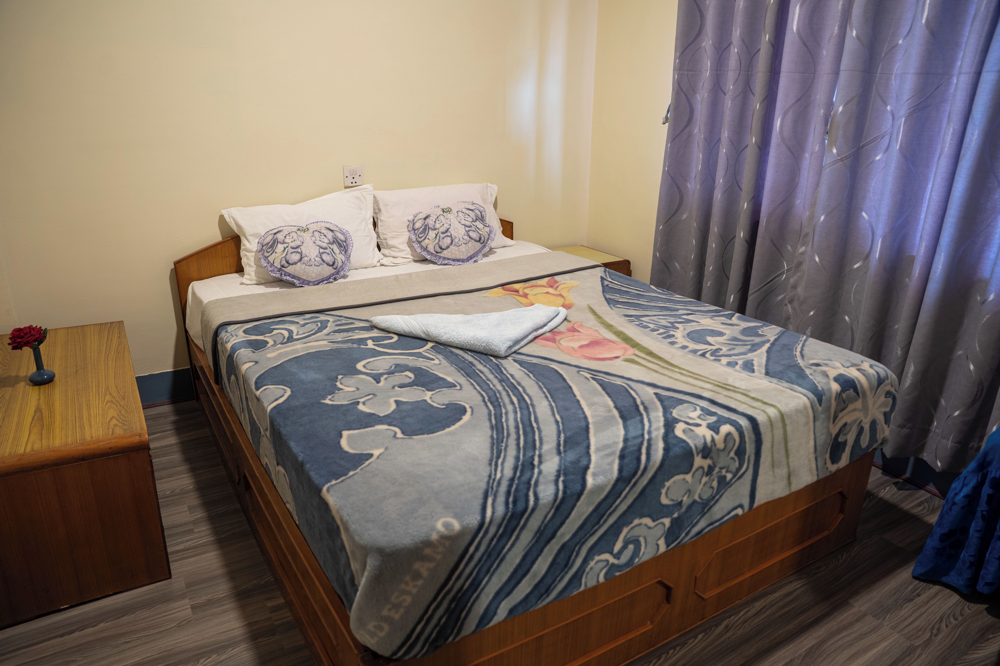 A neatly made bed with patterned bedding, a wooden nightstand with a vase, and a window with curtains.