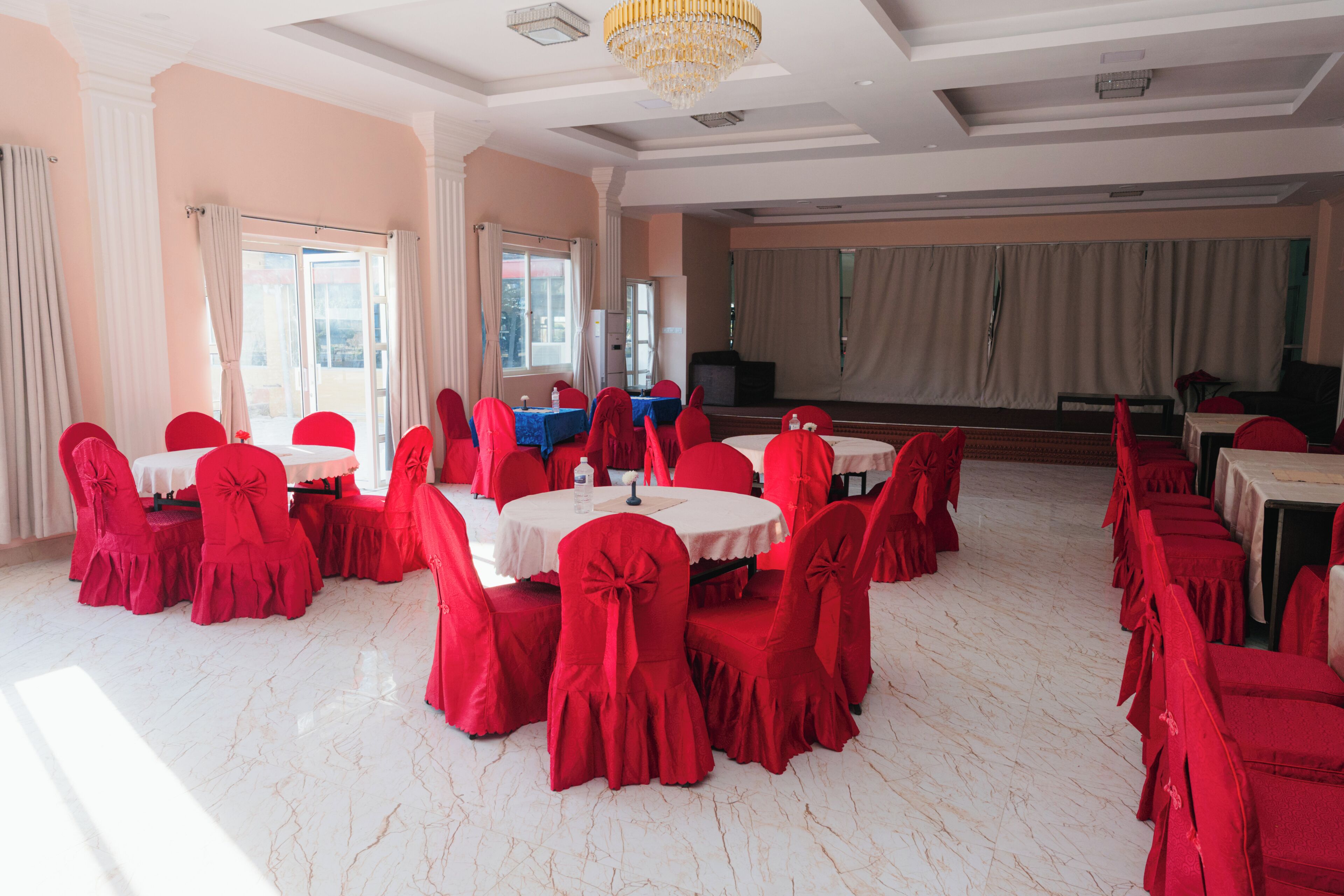 A banquet hall with round tables and red chairs, large windows, and a chandelier.