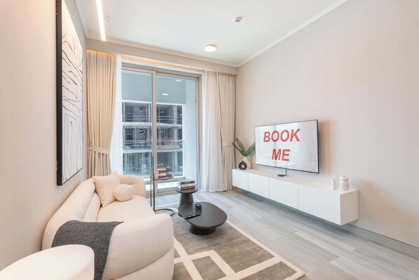 A modern living room with a sofa, a coffee table, a TV mounted on a wall, and a large window with curtains. - Free housekeeping visits during your stay - StayShort - Amazing 1 Bedroom in Concept 7 JVC Sleeps 2 (Jumeirah Village Circle)