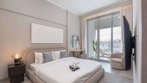 A modern hotel room with a large bed, a bedside table with a lamp, a flat-screen TV, a seating area with a chair, and a balcony with a view of the city. - StayShort - Amazing 1 Bedroom in Concept 7 JVC Sleeps 2 (Jumeirah Village Circle)