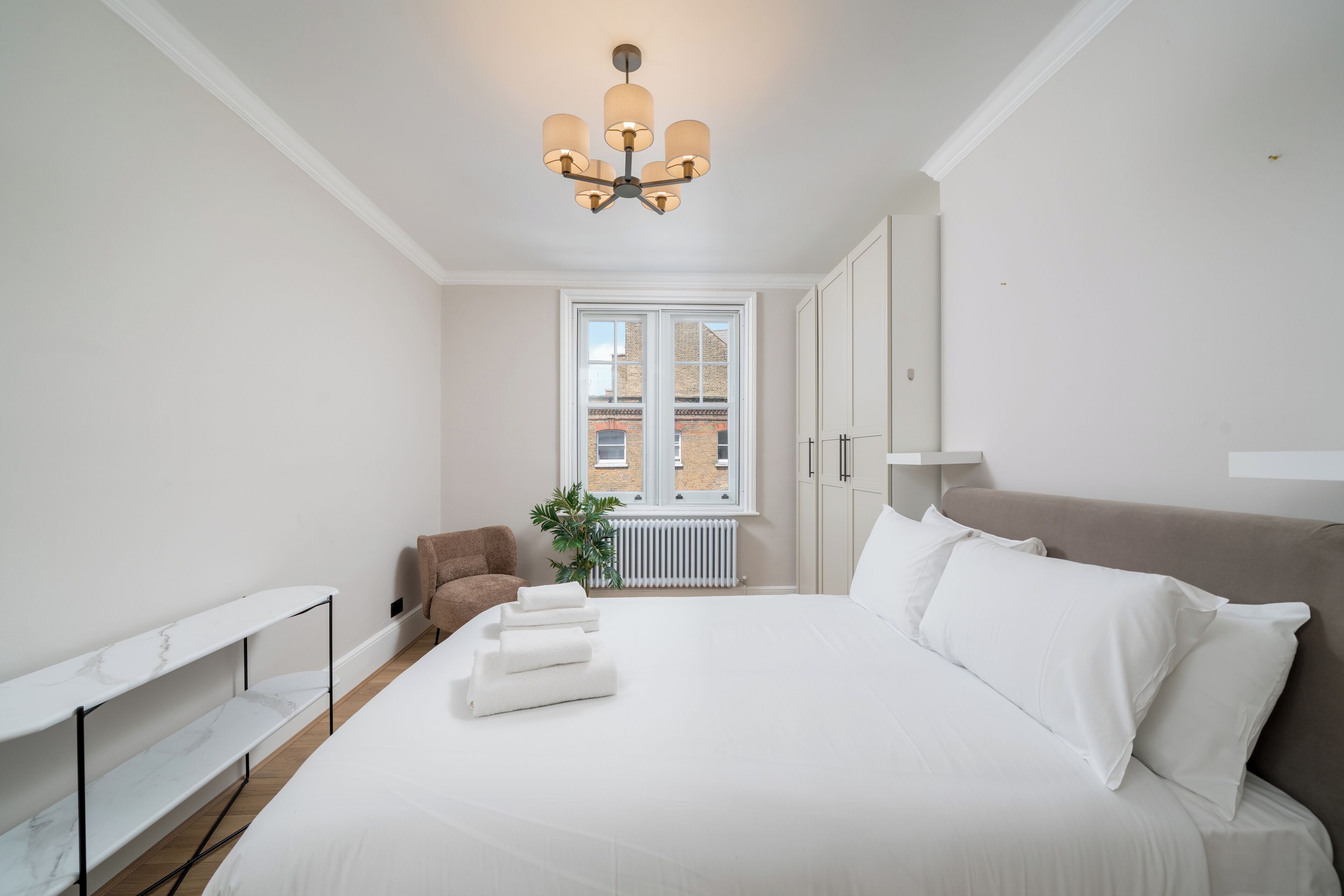 Un dormitorio moderno con una cama grande, un estante de mármol blanco, un sillón marrón y vistas a edificios a través de una ventana.