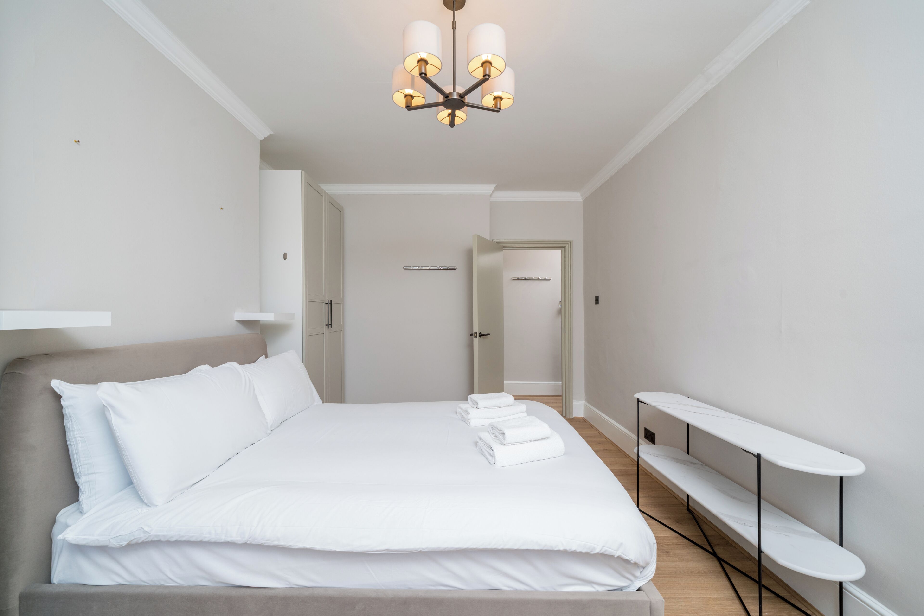 Quarto com cama, mesa de cabeceira, cadeira, luminária no teto e porta que dá para outro cômodo.
