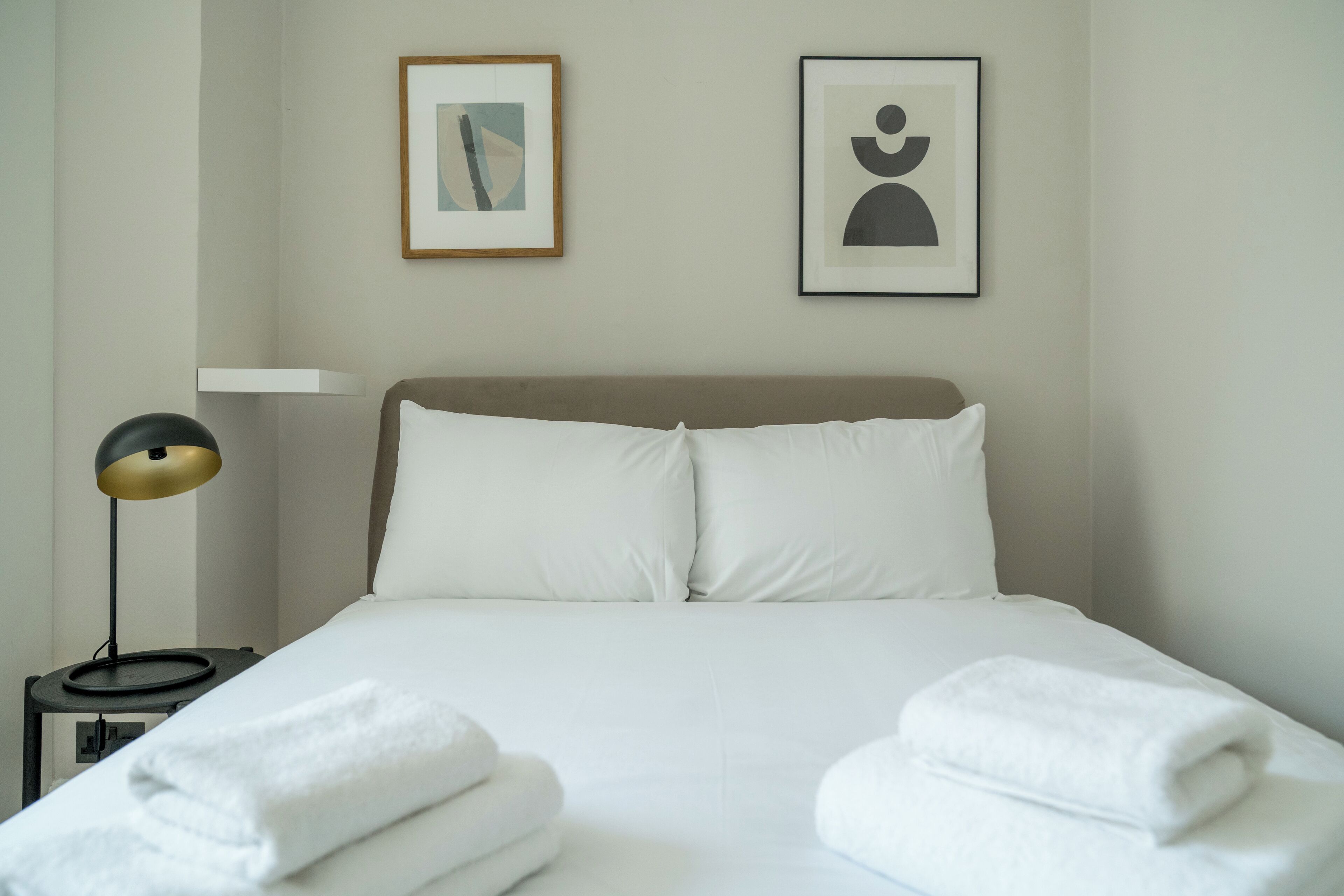 A neatly made bed with white linens and two rolled towels, a bedside lamp, and two framed artworks on the wall.