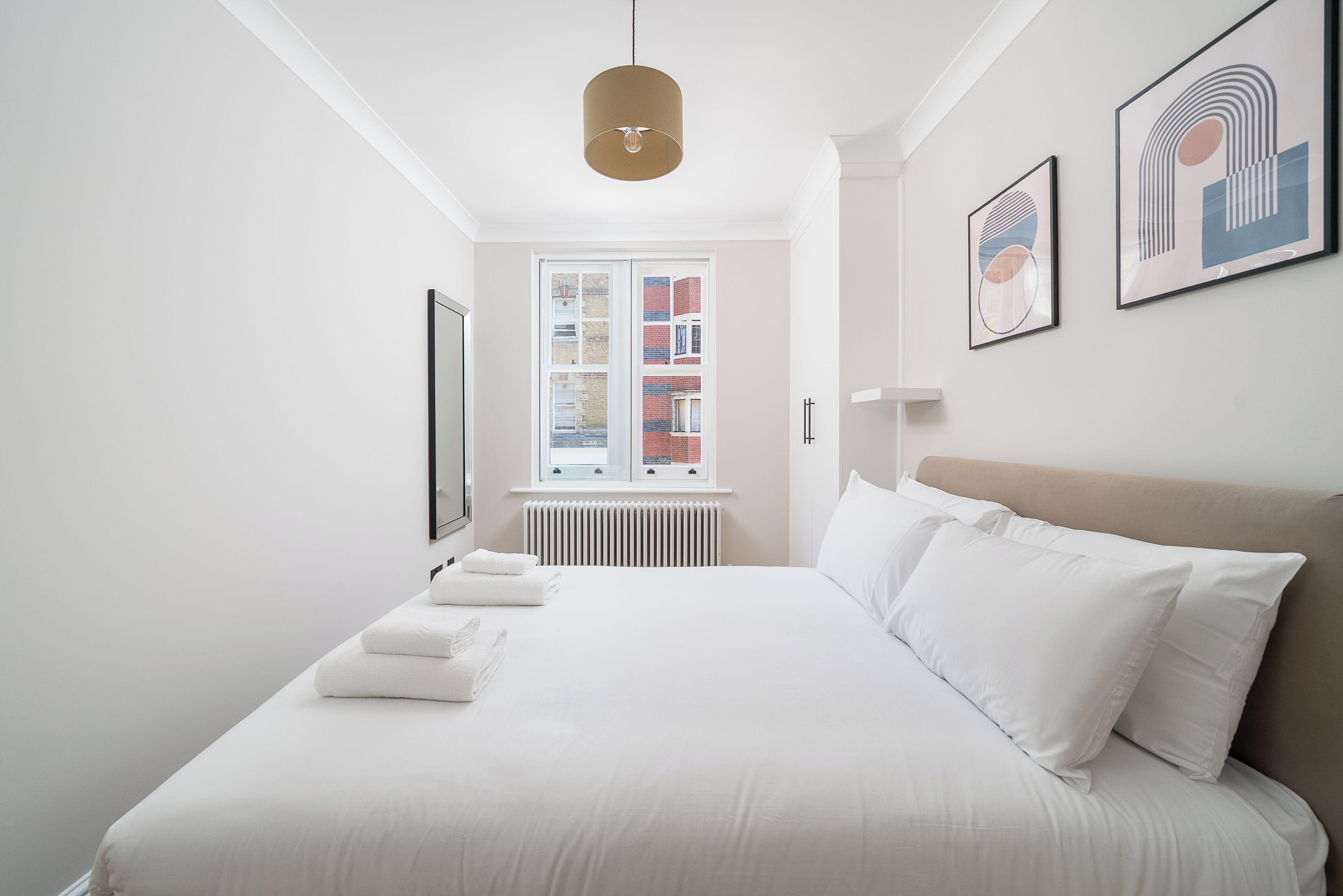 A neatly arranged hotel room with a large bed, a window with a view of buildings, and two framed artworks on the wall.