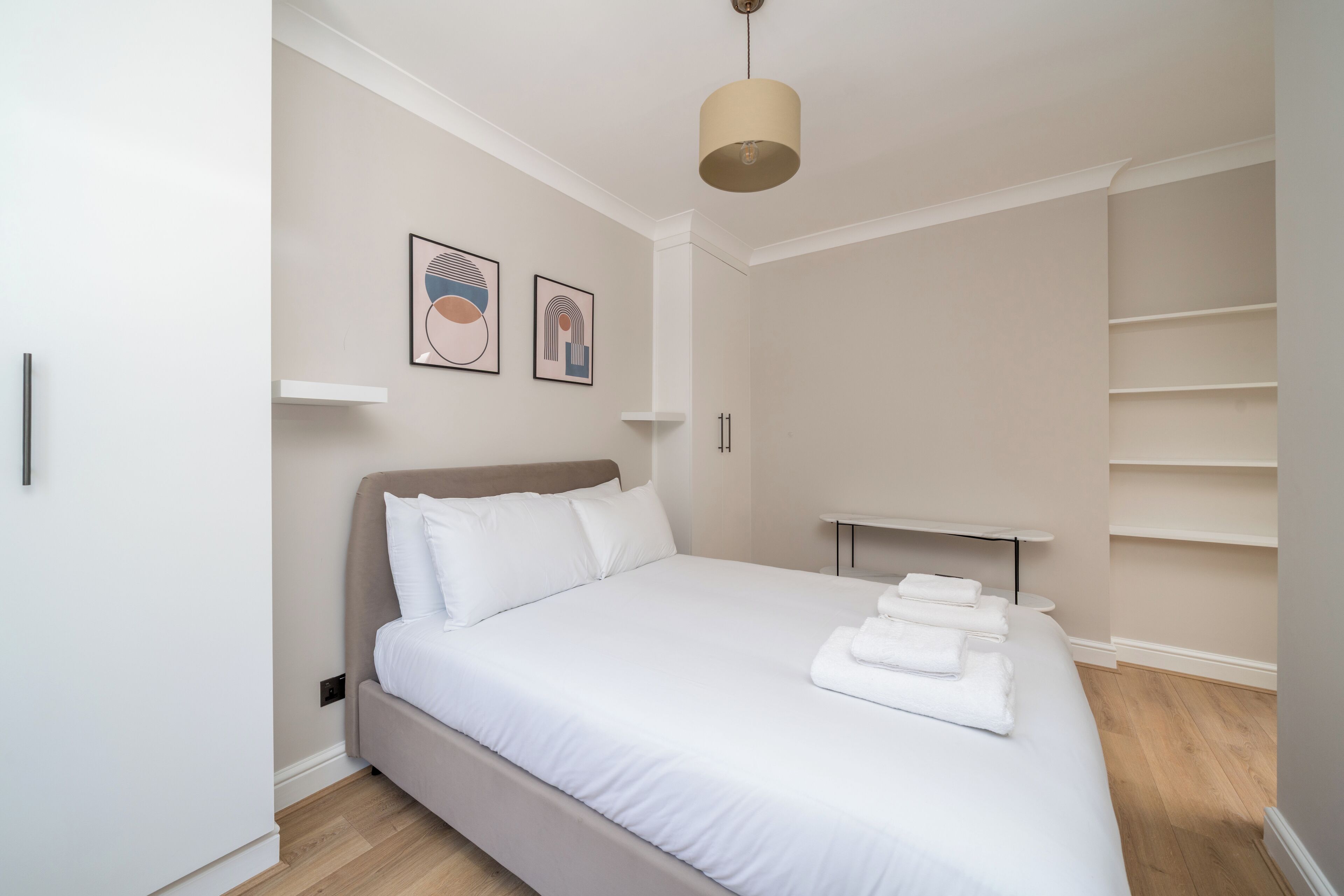 Ein ordentlich eingerichtetes Schlafzimmer mit einem großen Bett, einem Nachttisch, einer Pendelleuchte und zwei an der Wand befestigten Kunstwerken.