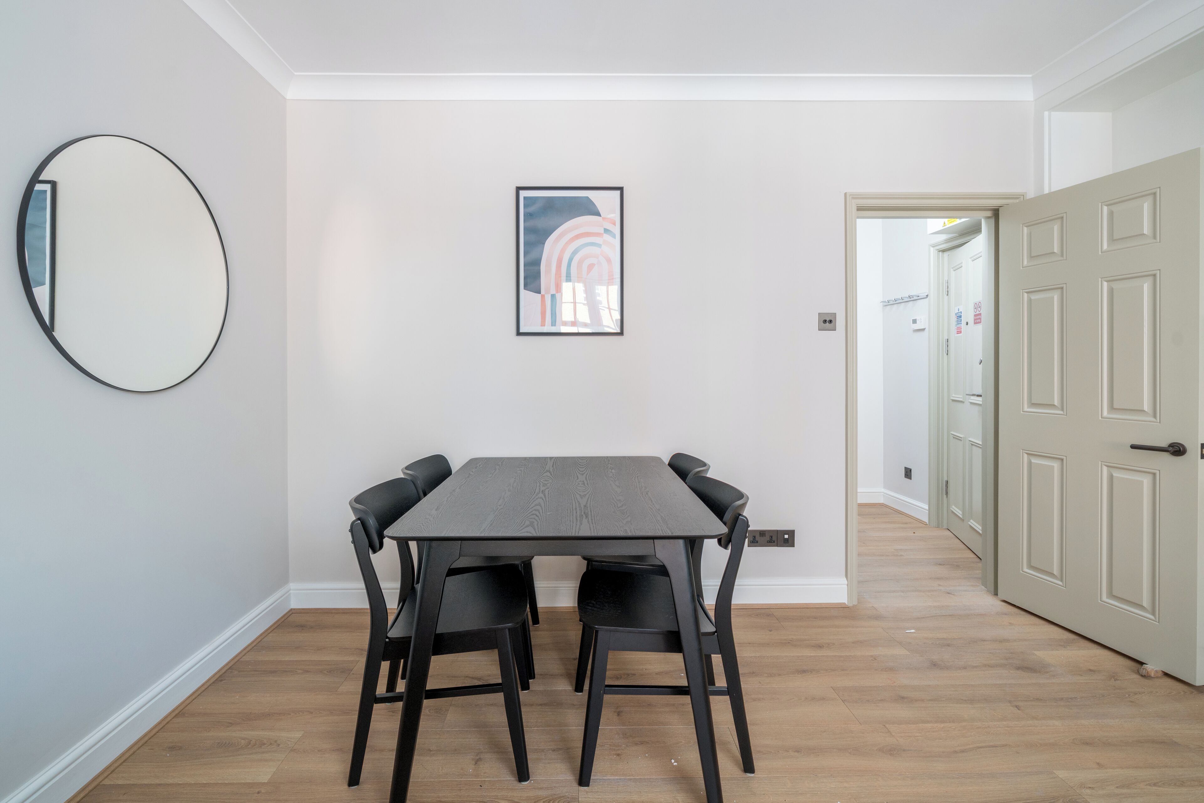A dining area with a rectangular table and four chairs, a round mirror, and a framed artwork on the wall.
