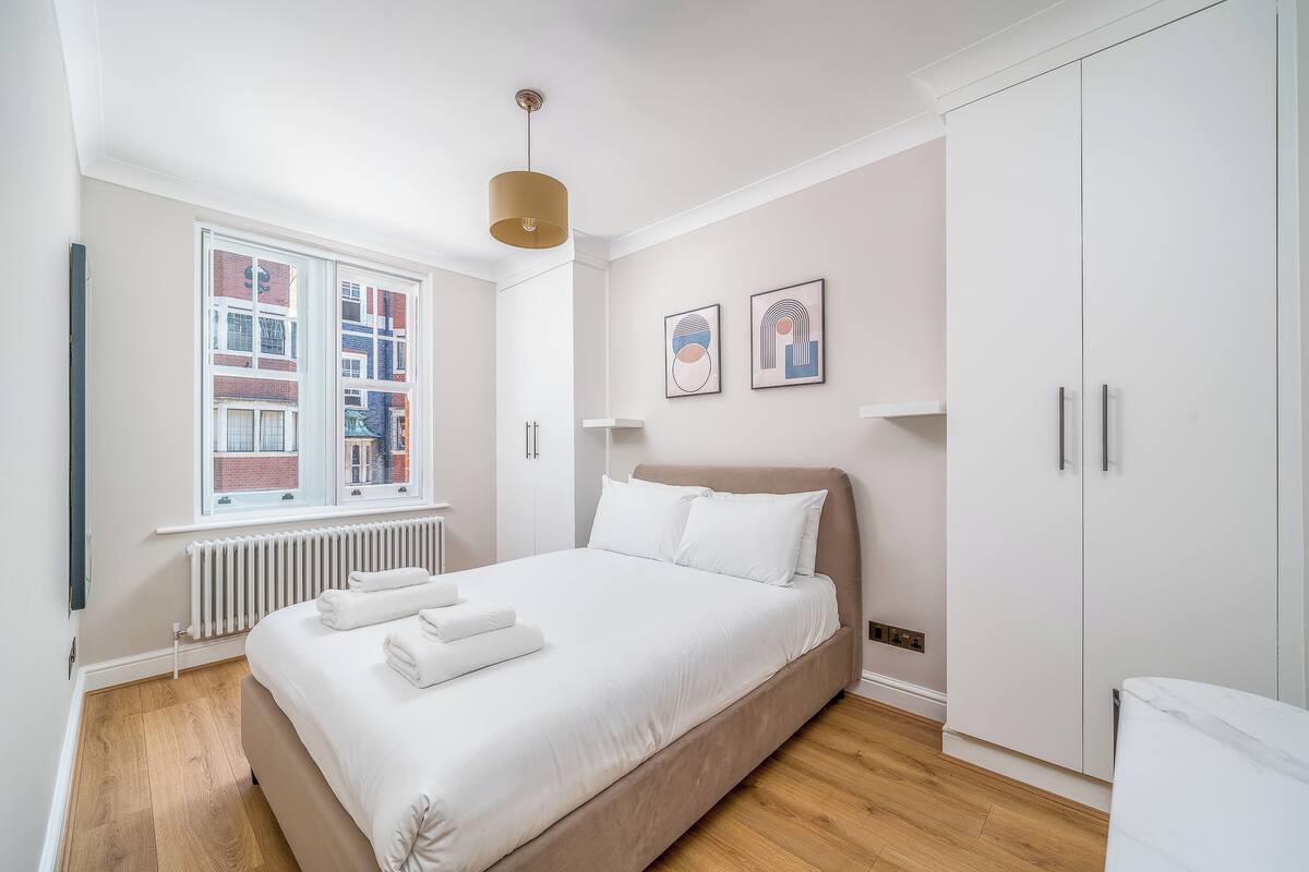 A modern bedroom with a large bed, white walls, a radiator, and a window with a view of buildings.