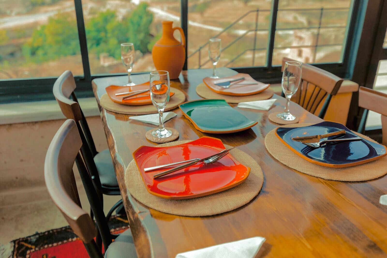 A dining table set with colorful plates, cutlery, and glasses, with a view of a landscape through large windows.