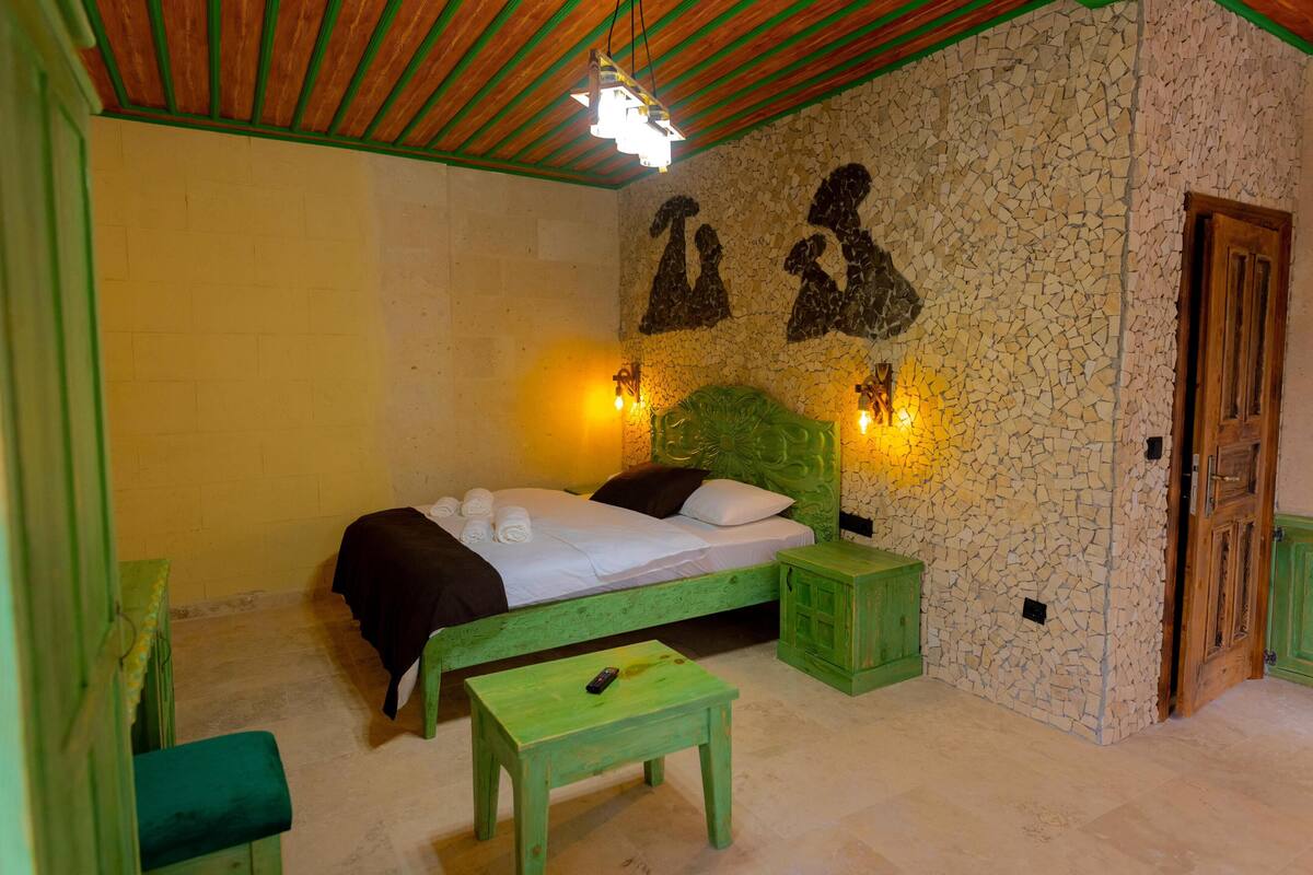 Ein Zimmer mit einem Bett, einem grünen Tisch, einer grünen Bank, einer Steinwand mit Hundeumrissen und einer Holztür.
