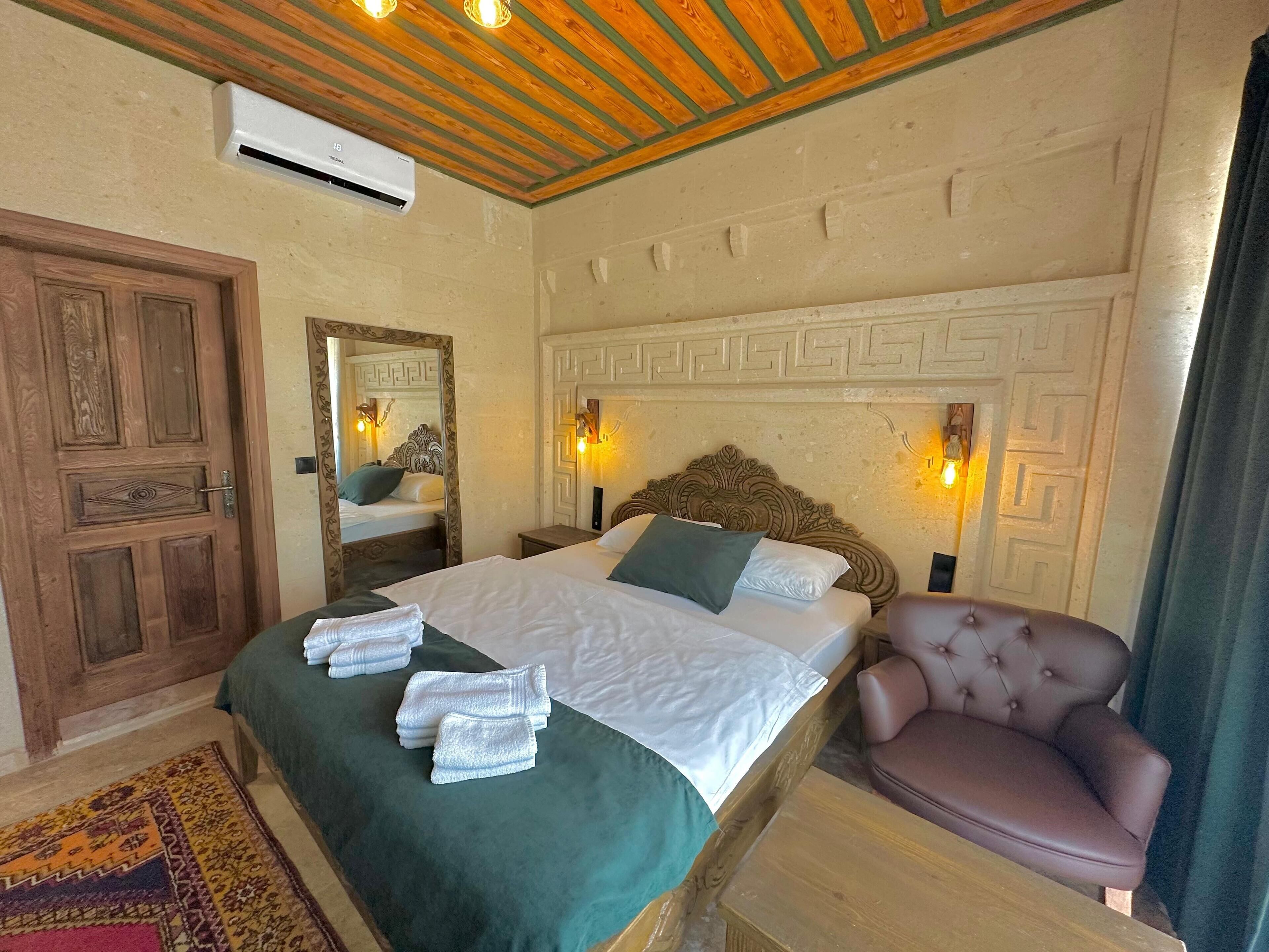 A bedroom with a large bed, a wooden door, a mirror, a headboard with wall lights, and a leather armchair.