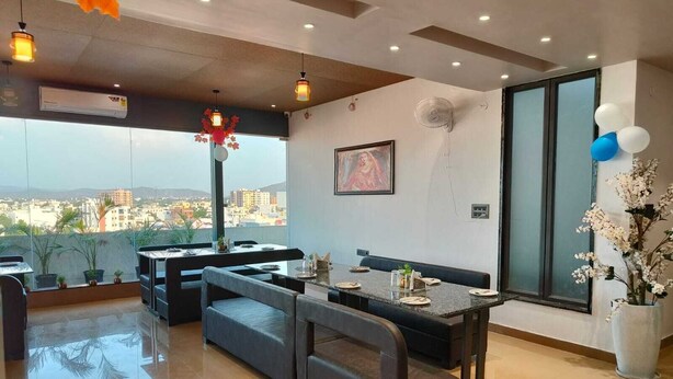 A modern dining area with a view of the city, a large window, and a ceiling fan. - Hotel Maple Residence (Udaipur)