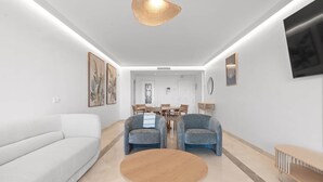 A modern living room with a white sofa, two armchairs, a round wooden coffee table, and a dining area with a wooden table and chairs.