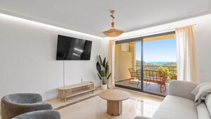 A modern living room with a flat-screen TV, a sofa, two armchairs, a coffee table, and a balcony with a view of the sea.