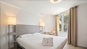 A hotel room with a large bed, two bedside lamps, a small table with a chair, and a view of greenery through the window.