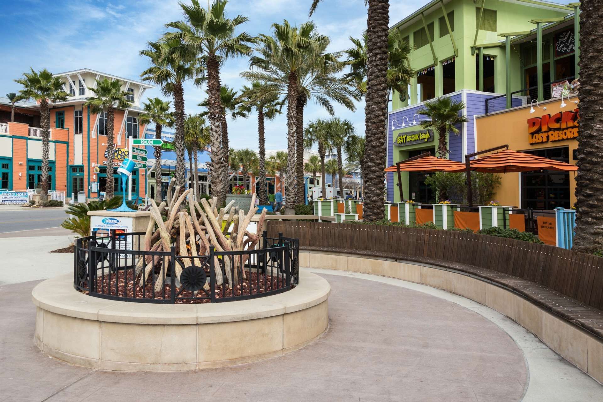Una plaza circular con una escultura de palmera, rodeada de más palmeras y diversos edificios comerciales.