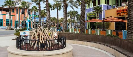 Una plaza circular con una escultura de palmera, rodeada de más palmeras y diversos edificios comerciales.