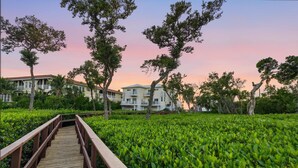 A wooden boardwalk leading to a building with multiple balconies, surrounded by lush greenery and trees.