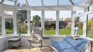 A conservatory with a cushioned chair, a small table with a tray, and a sofa with a blue blanket.