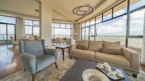 Ein modernes Wohnzimmer mit Sofa, Sessel und Couchtisch, großen Fenstern mit Stadtblick und einem Essbereich mit Stühlen und Tisch.