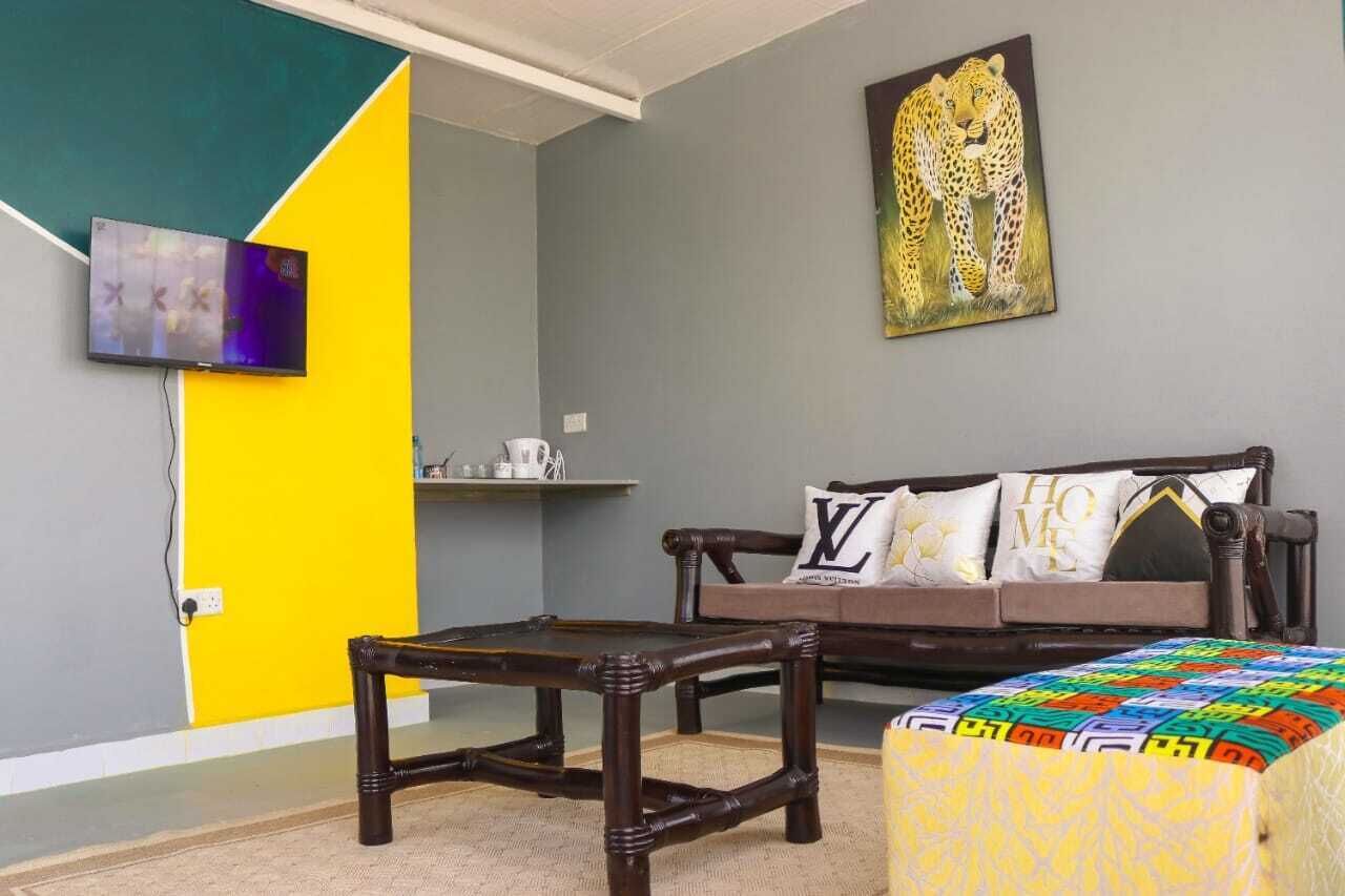Een kamer met een bank, een klein tafeltje, een kleurrijk poefje, een aan de muur bevestigde televisie en een schilderij van een luipaard.