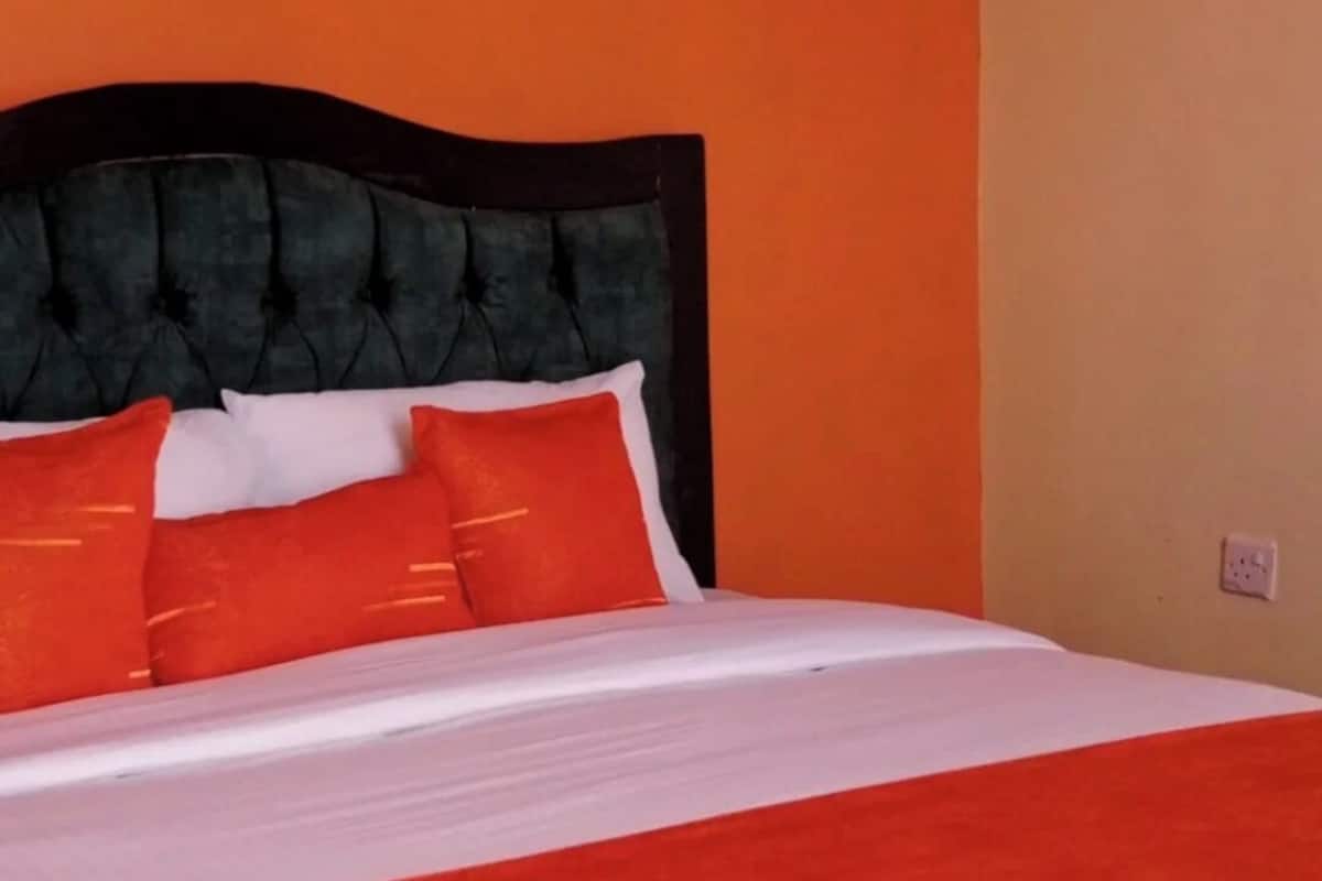 A bed with a white and orange bedspread, a black upholstered headboard, and multiple orange pillows.