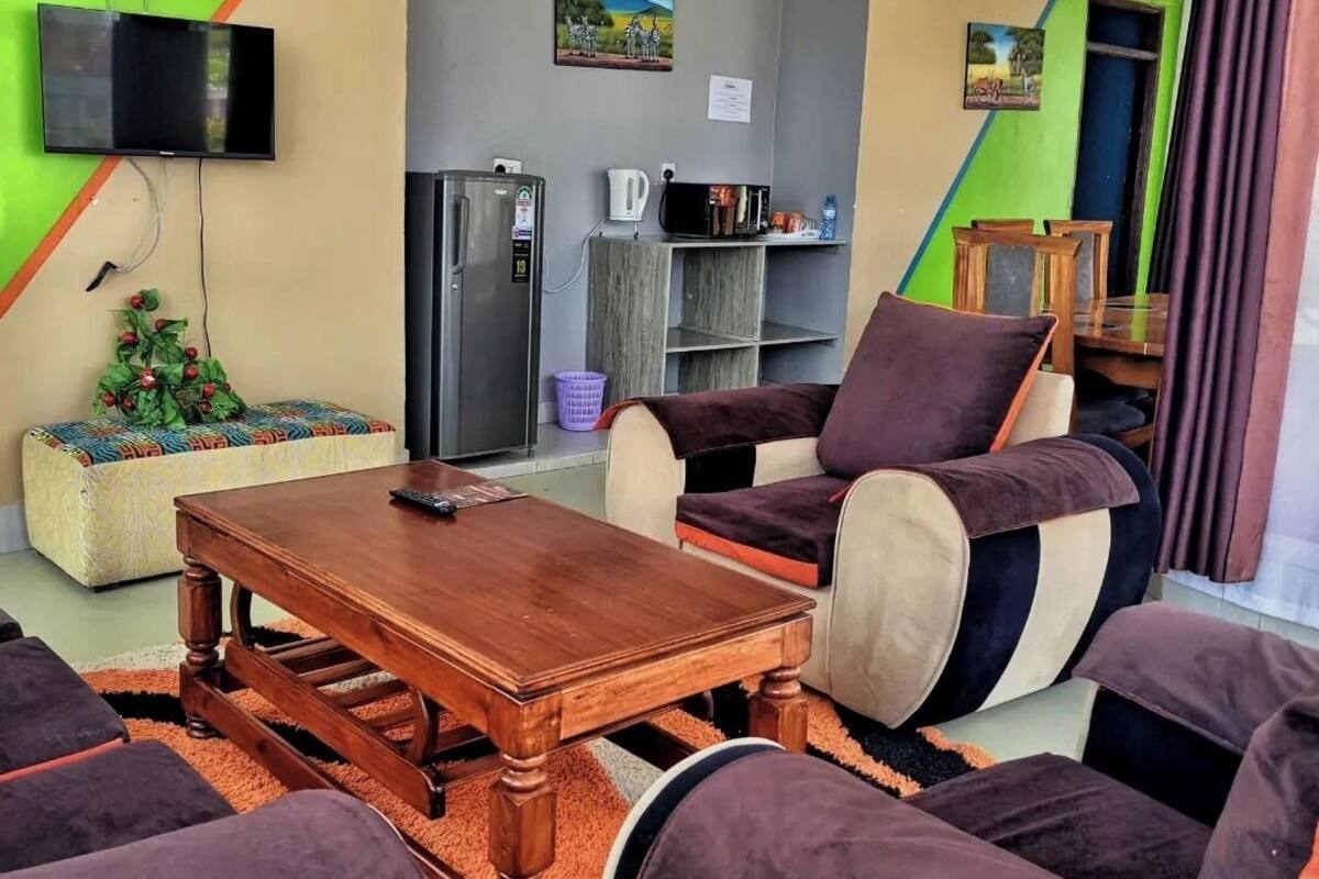 A living room with a wooden coffee table, cushioned seating, a flat-screen TV mounted on the wall, a refrigerator, and a microwave.