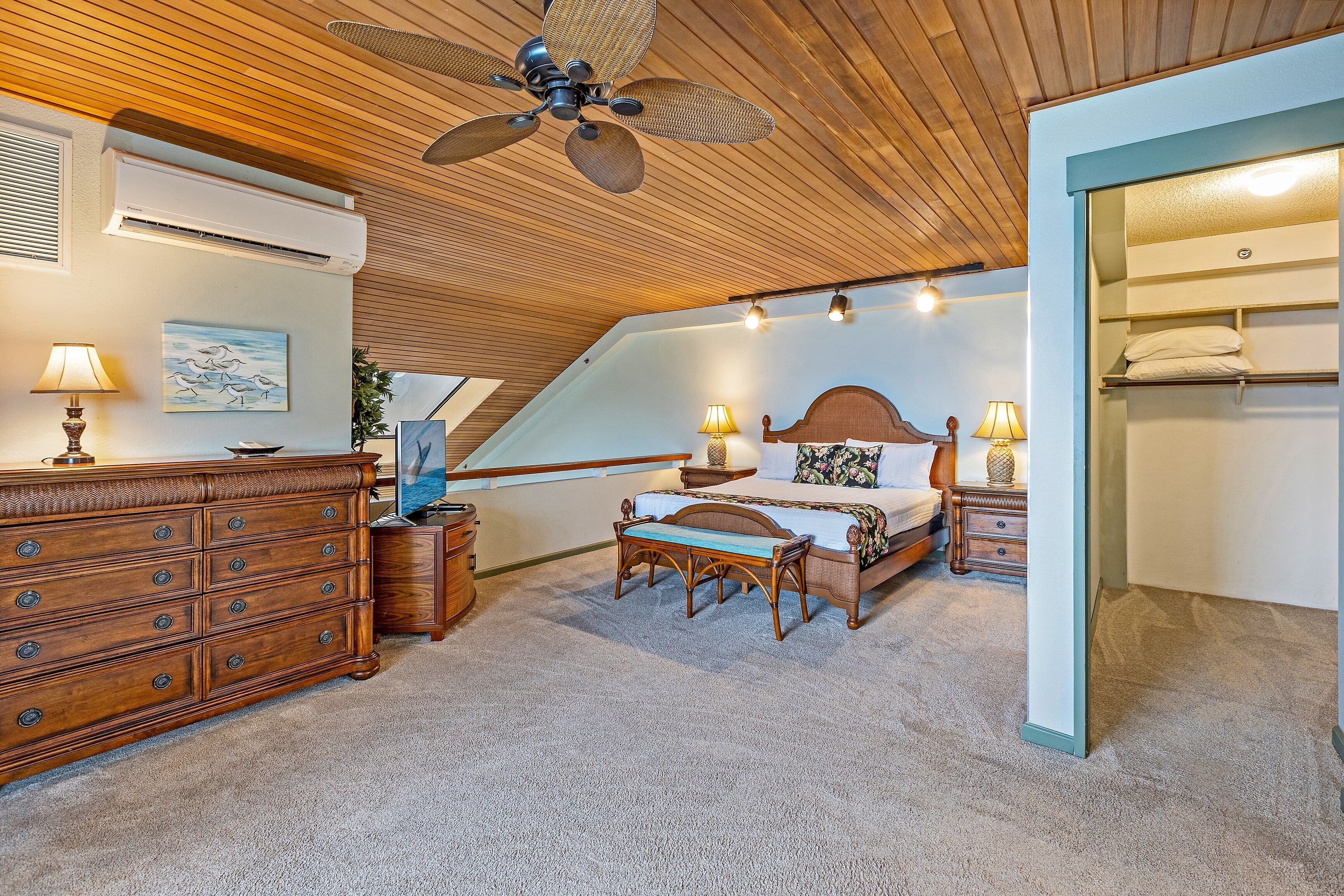 A bedroom with a sloped ceiling, wooden ceiling, a bed with a headboard, bedside tables with lamps, a dresser, and a bench.