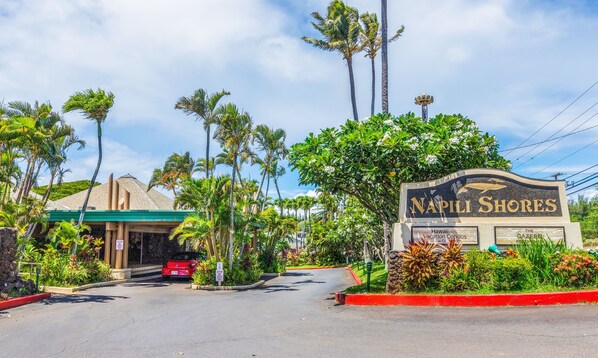 A sign for Napili Shores with a clear entrance, palm trees, and a building with a green roof.