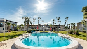 A swimming pool with a circular edge, surrounded by a fence, palm trees, and a building with balconies in the background.