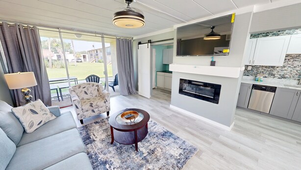 A modern living room with a sofa, armchair, and coffee table, a kitchen with white cabinets and a dishwasher, and a view of the outdoors through large windows.
