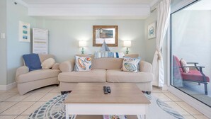 A living room with a sofa, two side tables, a coffee table, and a large window with curtains. - Tradewinds 102 1 Bedroom Condo (Orange Beach)