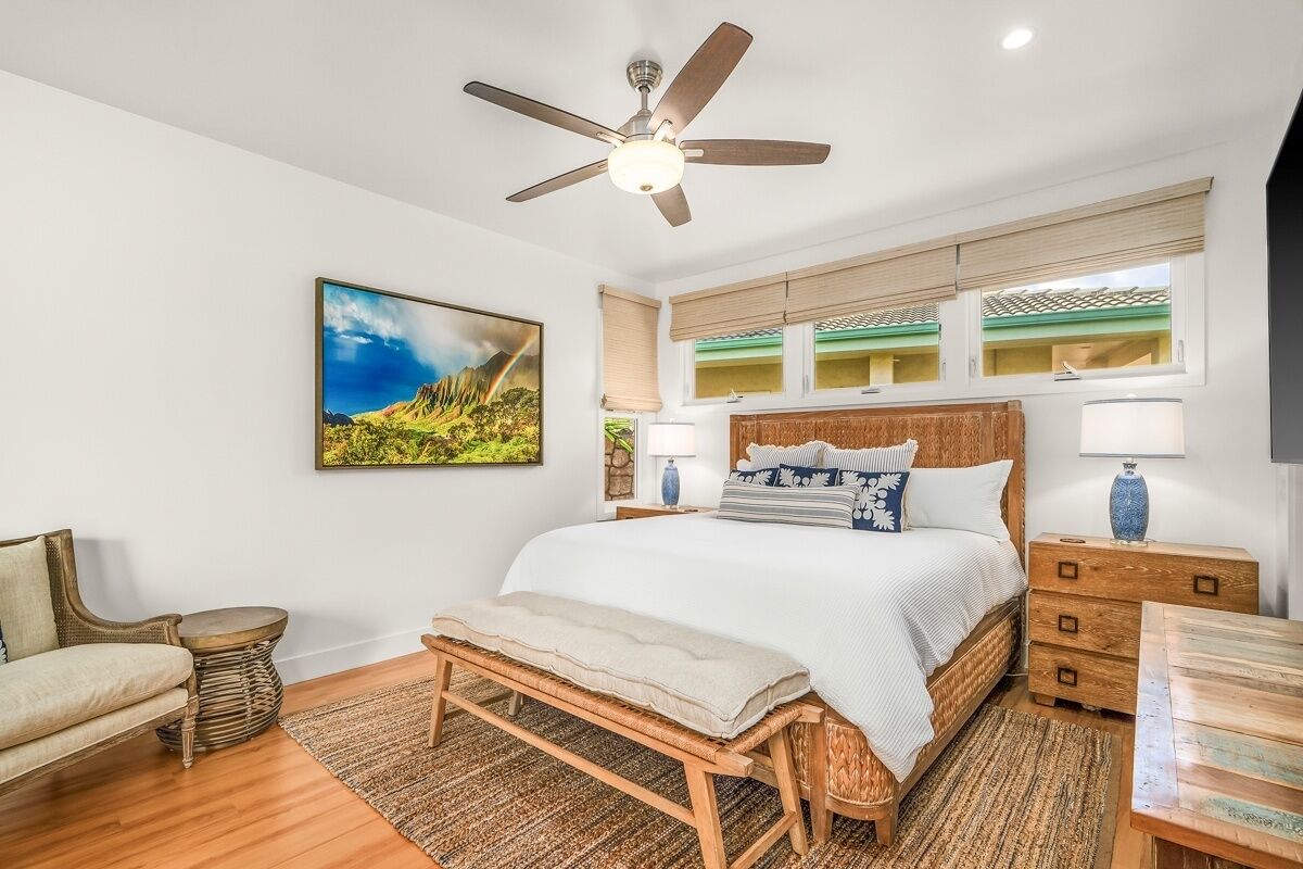 Een slaapkamer met een bed, een plafondventilator, een nachtkastje, een lamp, een stoel en een schilderij aan de muur.