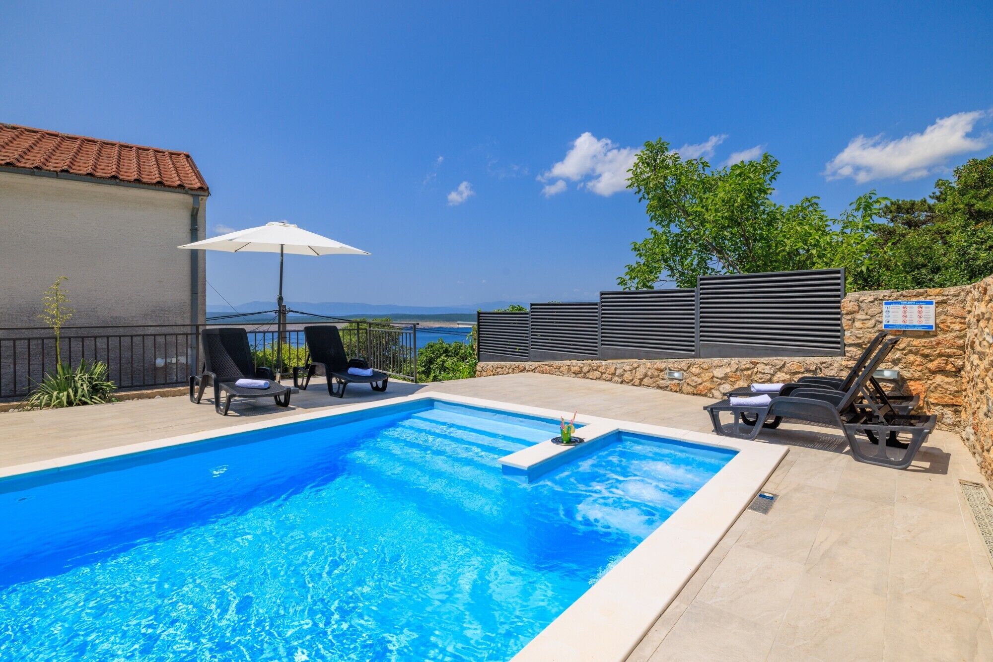 En pool med solstolar och parasoller, en stenmur och utsikt över havet.