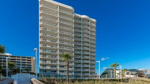 A tall, multi-story hotel with balconies, located near a beach with palm trees and a wooden walkway. - Tradewinds 102 1 Bedroom Condo (Orange Beach)