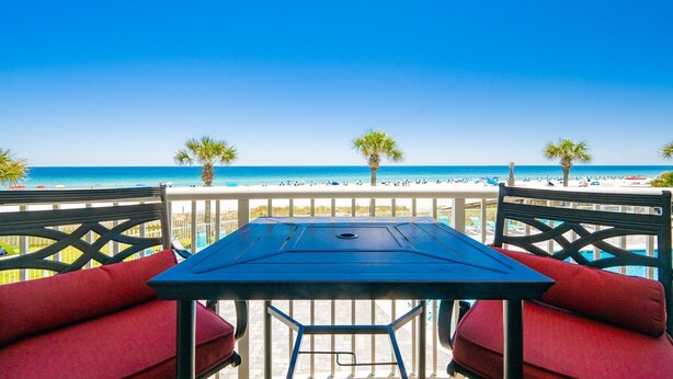 A balcony with a table and chairs, overlooking a beach with palm trees and the ocean. - Tradewinds 102 1 Bedroom Condo (Orange Beach)