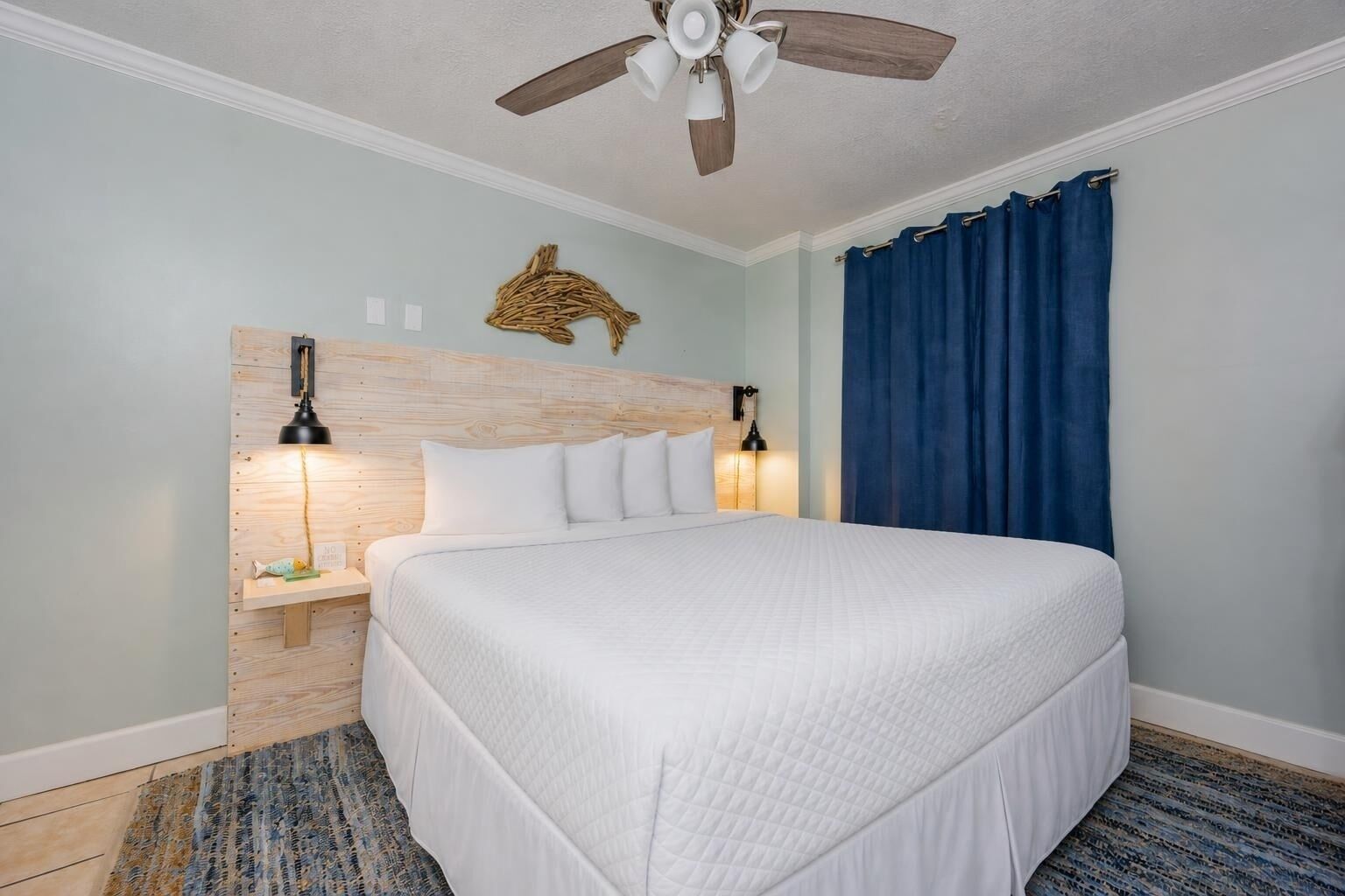 Habitación de hotel con una cama grande, un ventilador de techo, una lámpara de pared y una cortina azul.