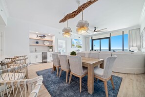 Une cuisine moderne avec des meubles blancs, un coin repas doté d’une table en bois et de chaises en osier, et une grande fenêtre offrant une vue sur le ciel.