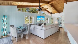 Amplia sala de estar con techo de madera, una mesa de comedor con sillas, un sofá blanco y un televisor mostrando una escena de playa.