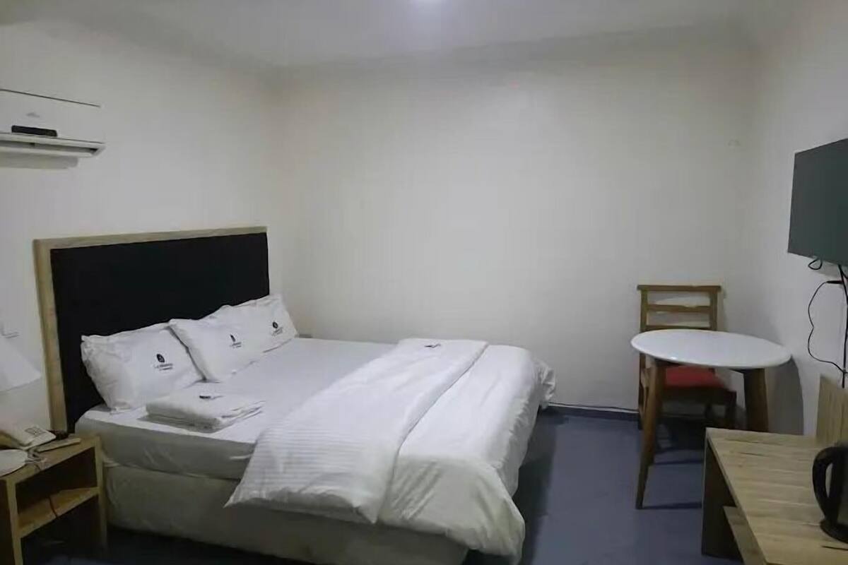 Un lit simple avec une literie blanche, une table de chevet avec une lampe, une petite table ronde avec une chaise, et un téléviseur fixé au mur.