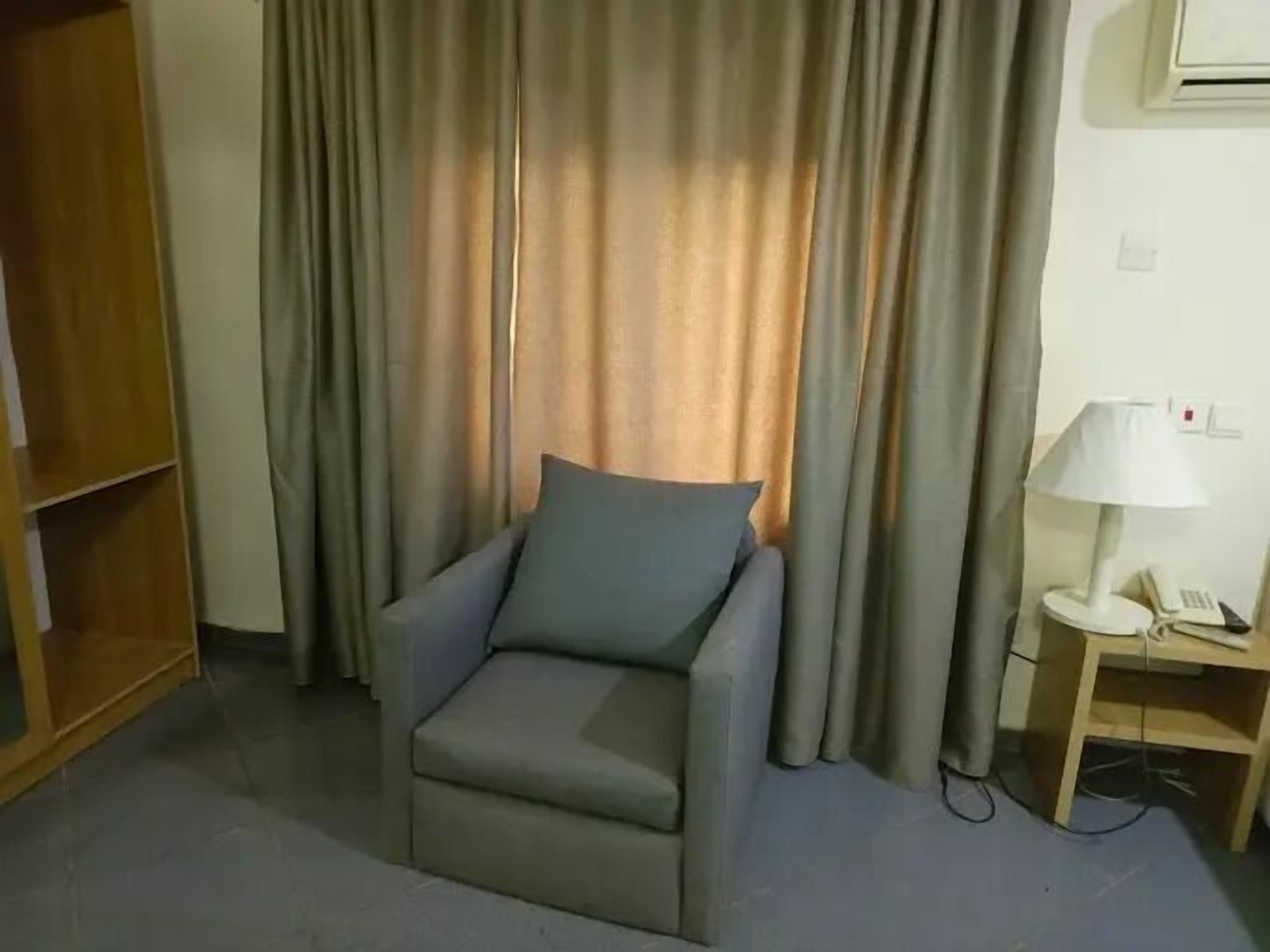 A small, single-occupancy room with a grey armchair, a wooden nightstand with a lamp, and a wardrobe.