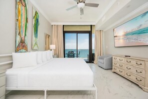 A hotel room with a large bed, a ceiling fan, a balcony with a view of the ocean, and a painting of fish on the wall.