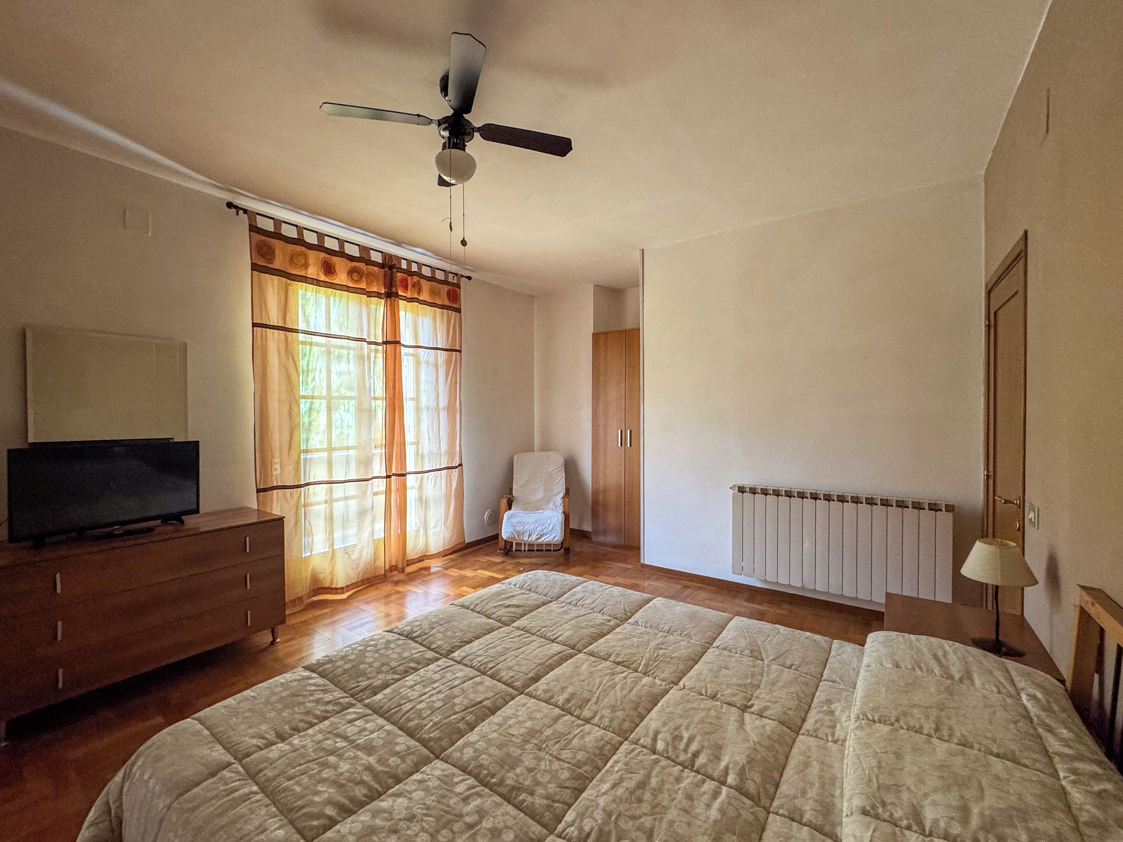 A bedroom with a bed, a TV on a dresser, a chair, a wardrobe, and a ceiling fan.