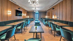 A hotel dining area with multiple tables and chairs, a central chandelier, and a television screen on the wall.