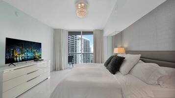 A modern hotel room with a large bed, a flat-screen TV on a white dresser, a balcony with a city view, and a chandelier.