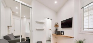 Uma sala de estar moderna com sofá, televisão fixada na parede e uma pequena cozinha com máquina de café.