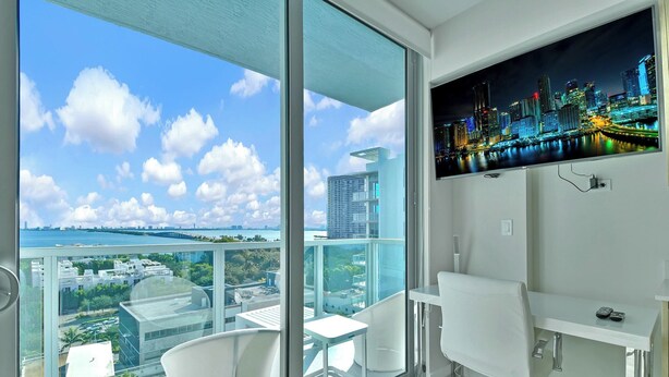 A modern room with a large window, a desk, a chair, and a mounted TV displaying a cityscape. - Design District Studio · City View · Free Parking (Miami)