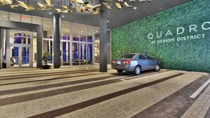 A modern building entrance with a car parked in front, large pillars, and a green wall with the text "QUADRO AT DESIGN DISTRICT". - Design District Studio · City View · Free Parking (Miami)