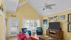 Ein Wohnzimmer mit rotem Sofa, blauen Sesseln, einem Holztisch, einem Fernseher auf einem Holzschrank und einem Deckenventilator.