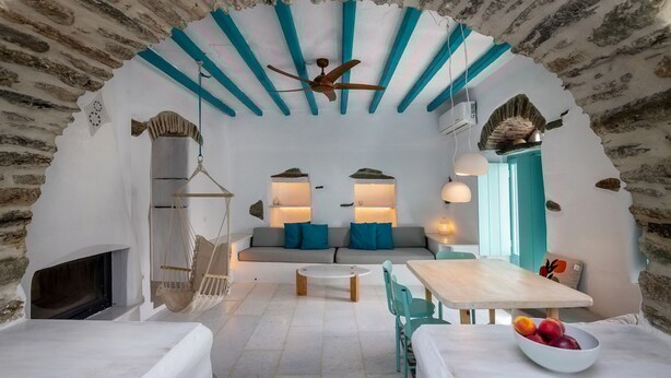 A cozy room with a stone arch, a ceiling fan, a sofa with cushions, a hanging chair, a table with chairs, and a bowl of fruit. - Melina House Platia Tinos (Tinos)